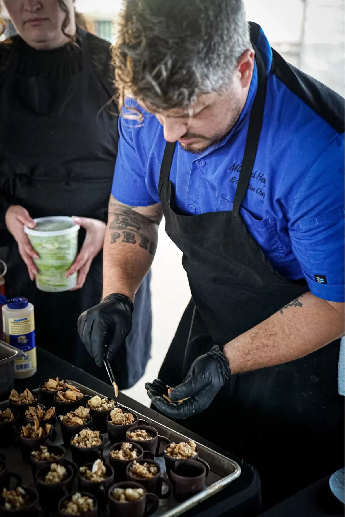 Chef in a blue jacket plating small bites onto a tray of dark cups