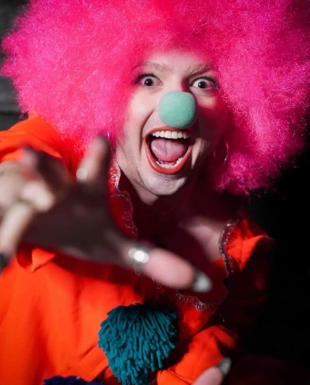 Performer in a pink clown wig and costume reaching toward the camera