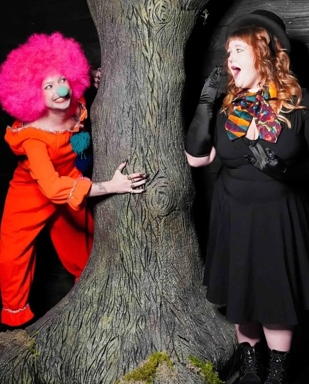 Costumed clown peeking around a tree at a startled performer in black