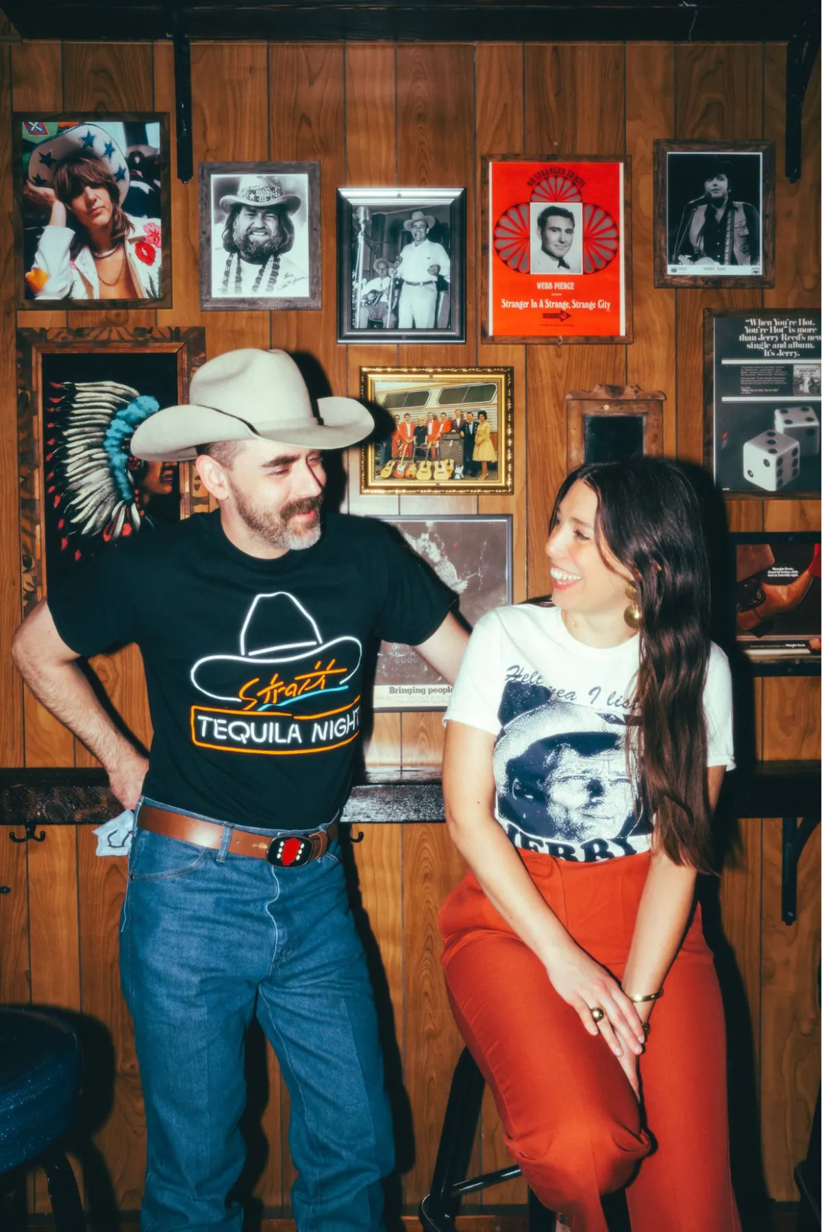 Two people in western wear posing in a honky tonk with framed photos on the wall